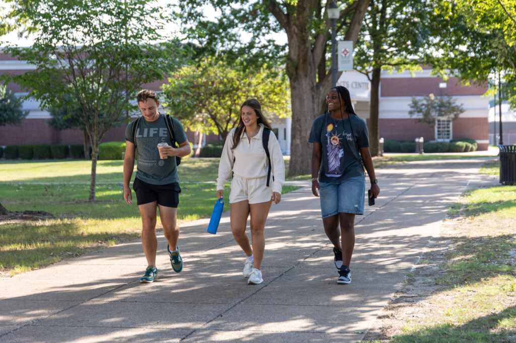 Three college students walk together on a sunny campus pathway, carrying backpacks and water bottles, with trees and a building visible in the background.