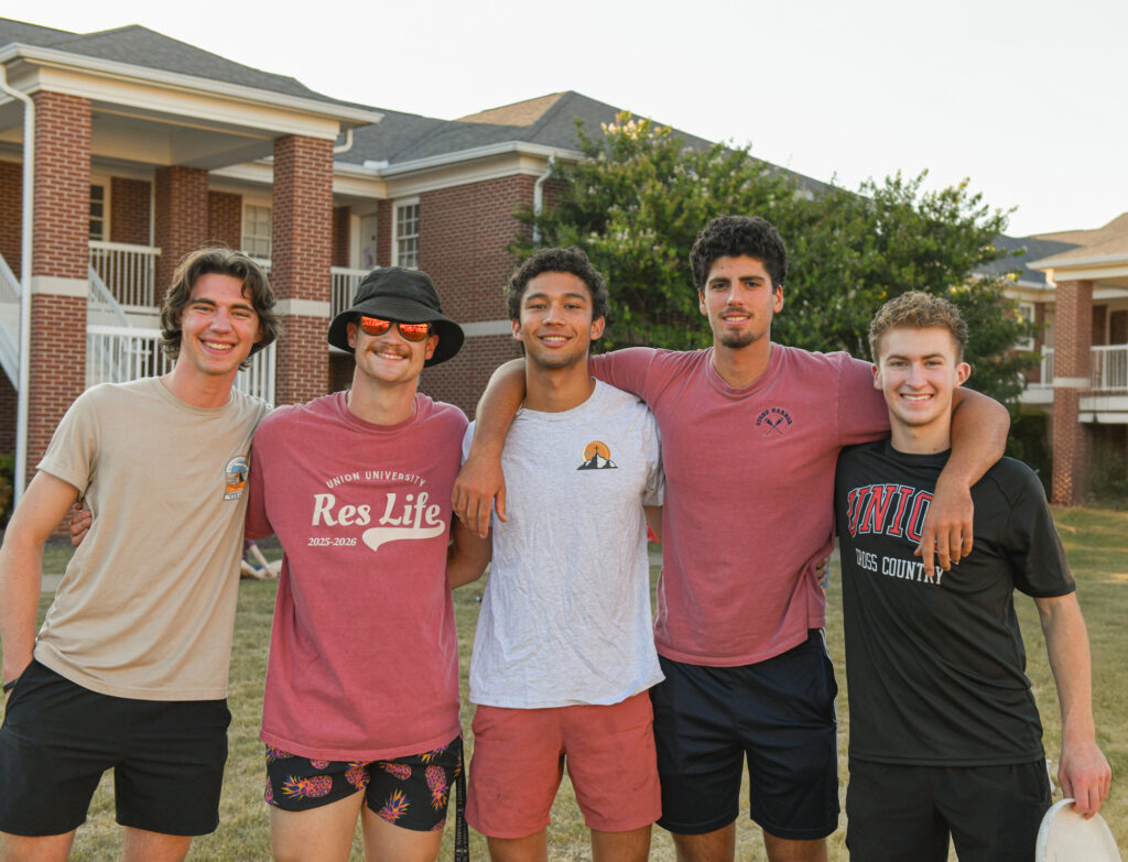 Five young men stand together outside, smiling at the camera. They are casually dressed, with apartment-style brick buildings and greenery visible in the background.
