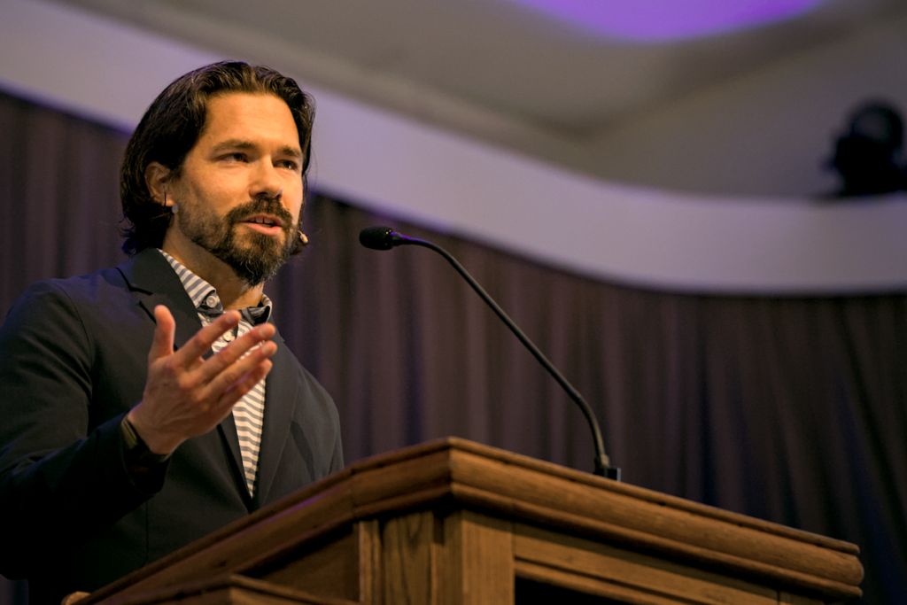 A man with long hair and a beard speaks at a podium on Biblical Studies, wearing a suit jacket and striped shirt, with a microphone headset and a curtained stage background.