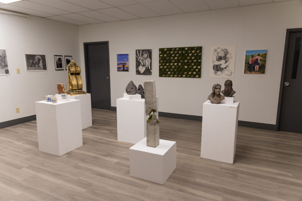 Art gallery room with striking sculpture displays on white pedestals and diverse artworks adorning white walls; the wood floor and ceiling tiles with fluorescent lighting create a modern showcase for art.