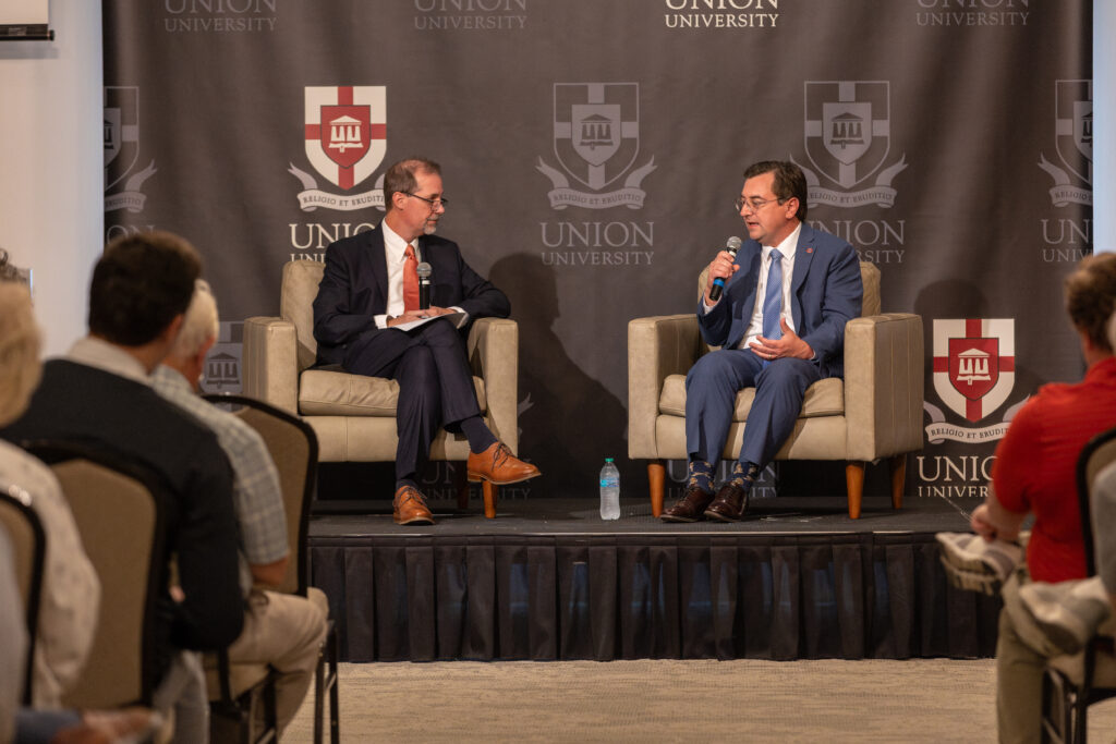 Two men in suits sit on stage in armchairs discussing ethics and public policy at a Union University event, with an audience seated in the foreground. A Union University banner is displayed behind them.