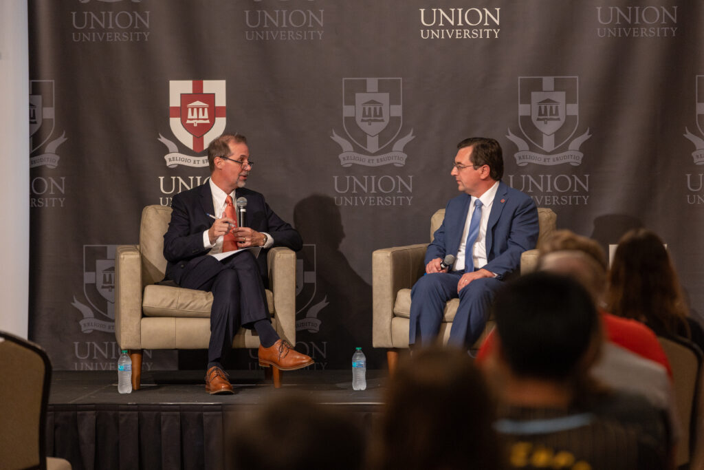 Two men in suits sit on stage in armchairs discussing Political Science at Union University. Both have bottled water at their feet, and an audience is seated in front of them. The university’s logo and banners are visible behind them.