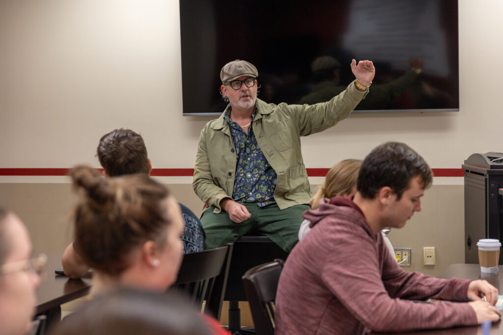 A man wearing glasses, a cap, and a green jacket sits on a desk in front of a classroom, gesturing with his right hand as several students studying Digital Media Communications listen to his storytelling.