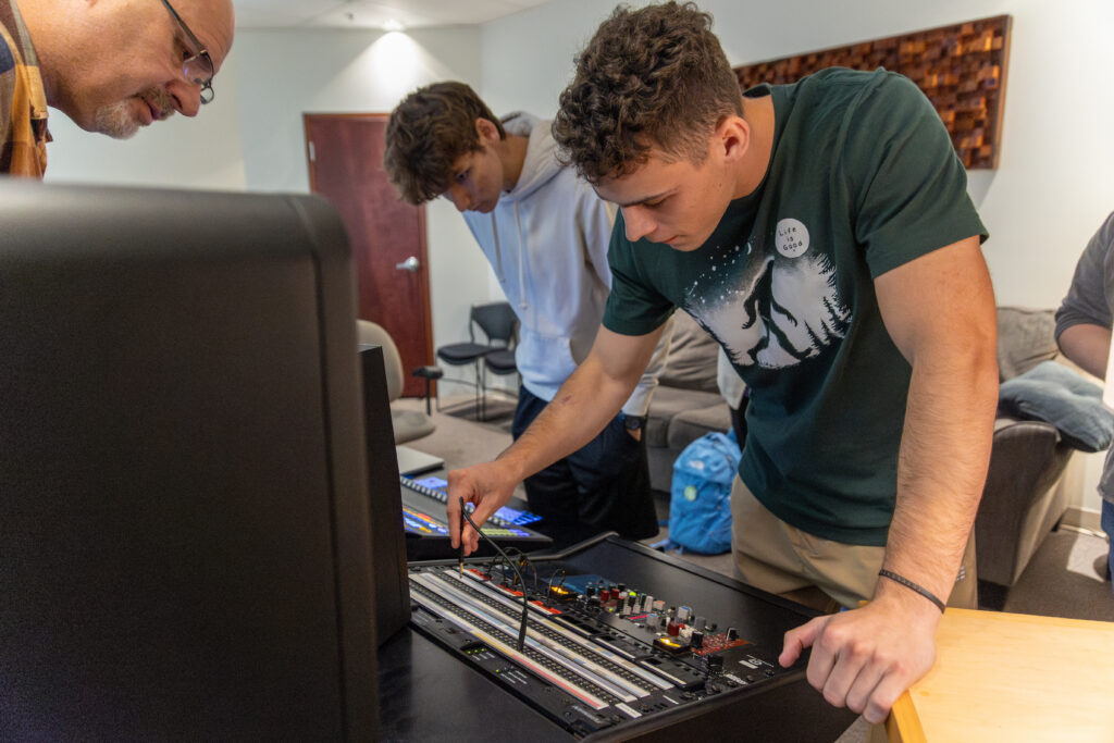 Three people stand around a sound mixing console in a recording studio, demonstrating recording engineering skills. One adjusts the controls while the others observe. A wall panel and seating are visible in the background.