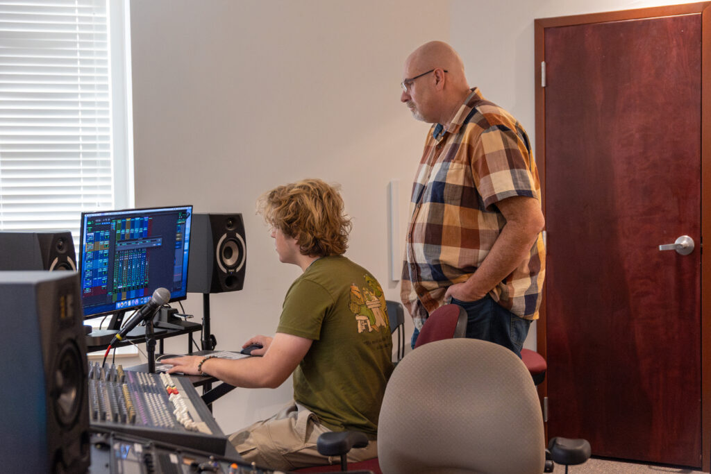 Two people in a sound studio work at a computer with audio editing software on the screen, collaborating on commercial music. One sits using the mouse while the other stands beside him, both focused on producing quality audio. Sound equipment fills the room.