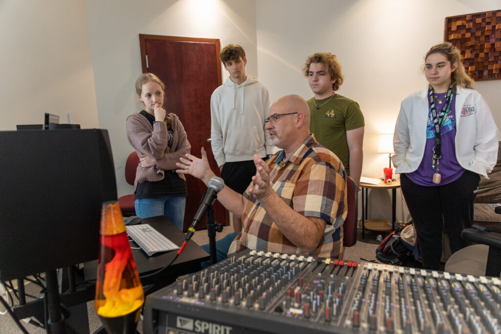 A man sits at a desk with audio equipment, speaking to four young adults in a studio setting. A lava lamp and microphone are visible on the desk, highlighting the engaging atmosphere of hands-on music education.
