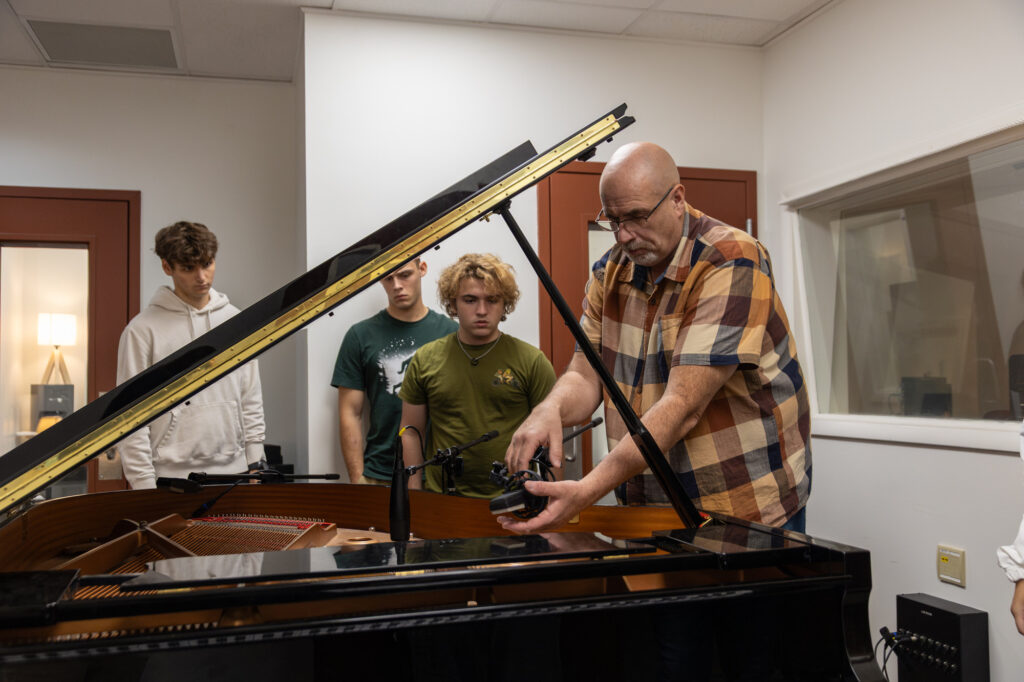 A man in a plaid shirt adjusts a microphone inside a grand piano while three young men, perhaps aspiring to earn a Music Technology degree, stand nearby watching in a recording studio.