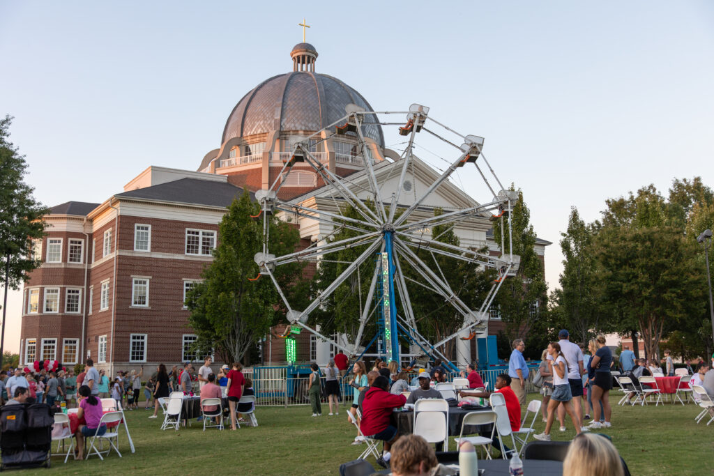 People gather on a lawn with tables and chairs in front of a Ferris wheel and a large domed building on Union's main campus in Jackson, Tennessee. Some ride the Ferris wheel while others are seated or standing in groups.