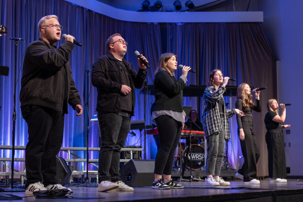 Six people from the Christian Ministry MMWL stand on stage singing into microphones. Dressed casually, they face the audience. Musical equipment and a drum set are visible in the background, with the stage lit in purple hues.