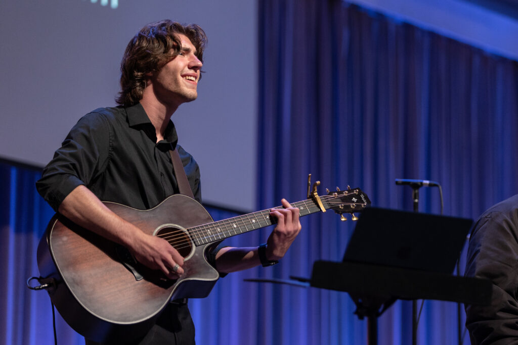 During a lively performance, a person in a black shirt plays an acoustic guitar on stage, smiling. Blue curtains and a microphone stand frame the scene, while a laptop sits on a stand in the foreground—perfect for commercial music settings.