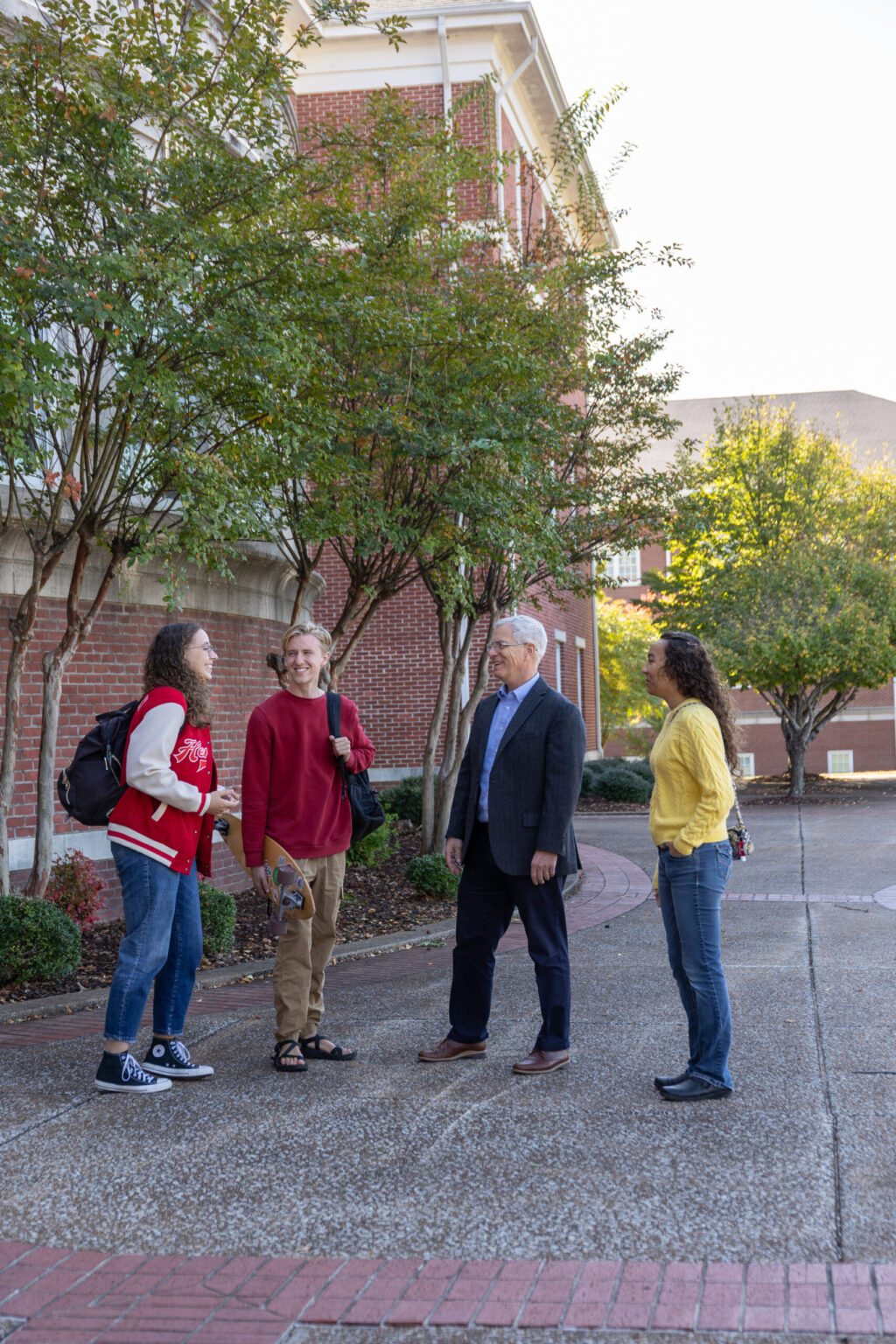 Four people stand outside on a brick pathway near a building, talking. Two appear to be students, carrying backpacks, while one man in a suit and a woman in a yellow sweater face them. Trees and shrubbery line the area.