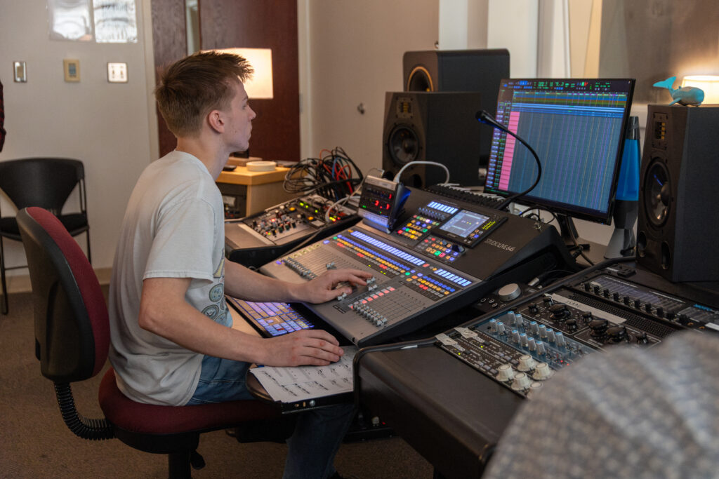 A person with a Music Technology Degree sits at a sound mixing console, adjusting controls and viewing audio editing software on a computer in a recording studio filled with audio equipment and speakers.