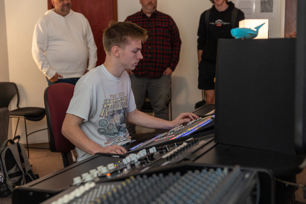 A young man sits at a large audio mixing console, adjusting controls, while three adults stand and watch in the background. The room appears to be a sound studio, highlighting the art of Recording Engineering and Music Producing.