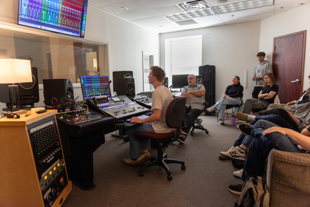 A person operates audio equipment at a mixing console in a studio recording session. Several people sit along the wall observing, surrounded by monitors and post-production technology throughout the room.