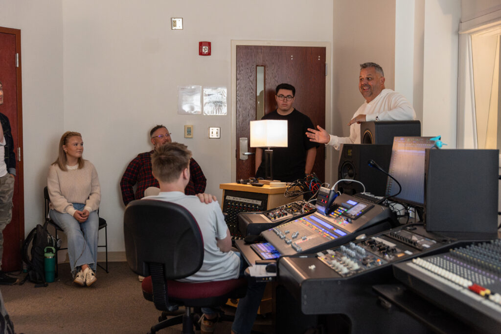 A group of people are gathered in a recording studio, engaging in Music and Communication Arts. Four are seated while one stands and gestures with his hand, surrounded by audio equipment and computer screens in the foreground.