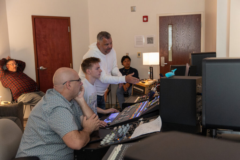Several people are in a sound control room; one man stands and points at a monitor while two others sit at a sound mixer, focused on music management and the screens. Two more people sit in the background observing.