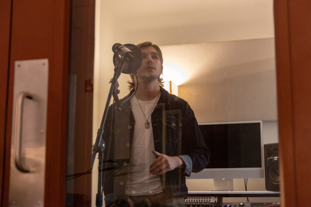 A person stands behind a glass door in a recording studio, singing or speaking into a microphone—perfect for songwriting or commercial music projects. A computer monitor, speakers, and soundproofing panels are visible in the background.