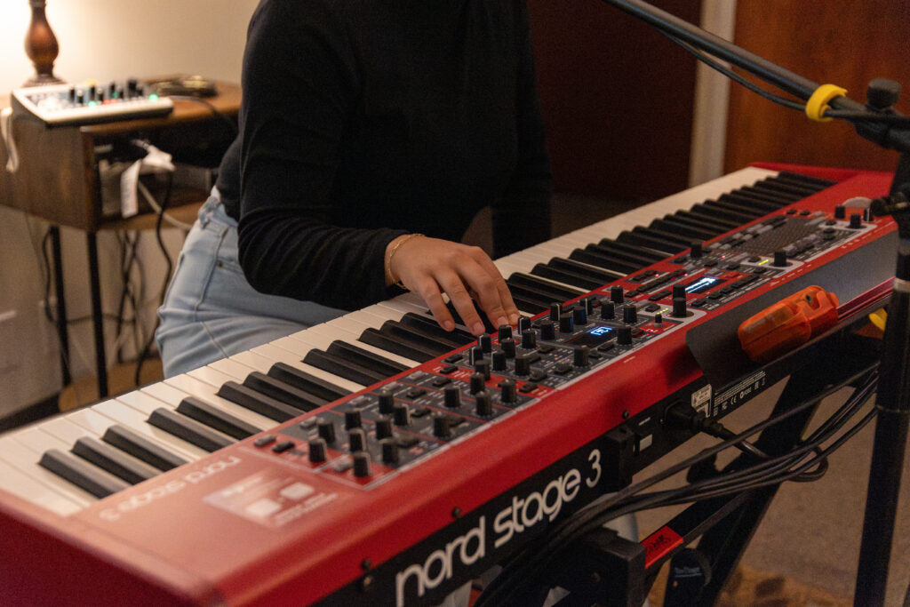 A person wearing a black long-sleeve shirt and light blue jeans plays a red Nord Stage 3 keyboard, showcasing their songwriting skills. Only the torso and arms are visible. A microphone and audio equipment are nearby.