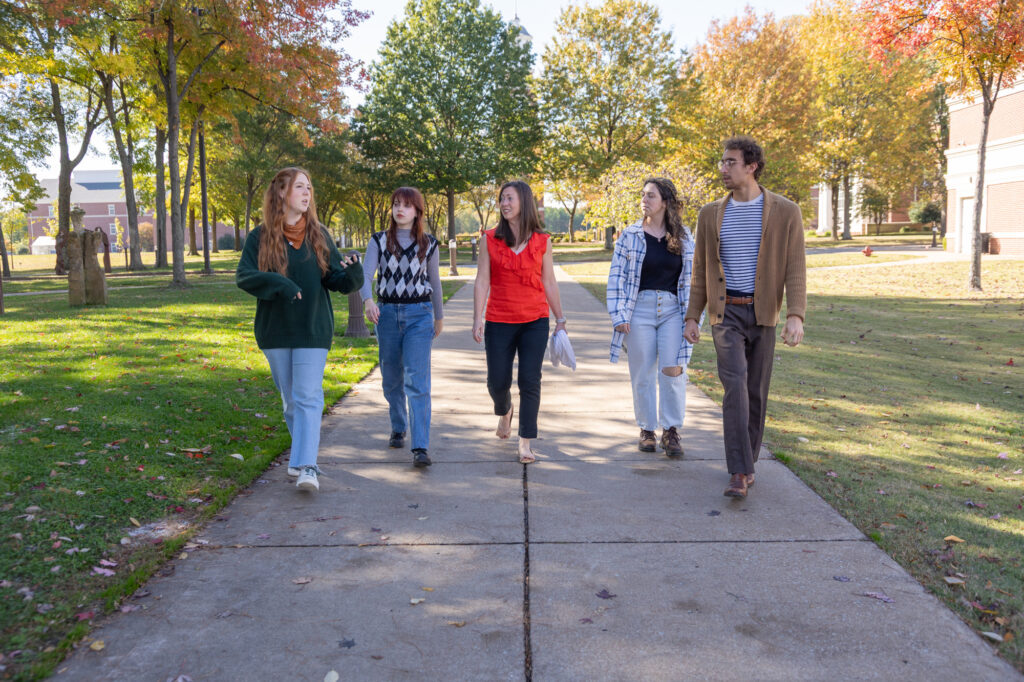 Five people, casually dressed, walk and talk together on a paved path in a park-like area with green grass and autumn trees. Sunlight filters through the leaves as the group discusses topics related to Honors Integrative Studies.