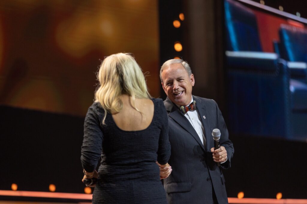 A man in a suit with a bow tie holds a microphone and smiles at a woman with long blonde hair, who faces away from the camera, on a stage with warm lighting and a large screen in the background.