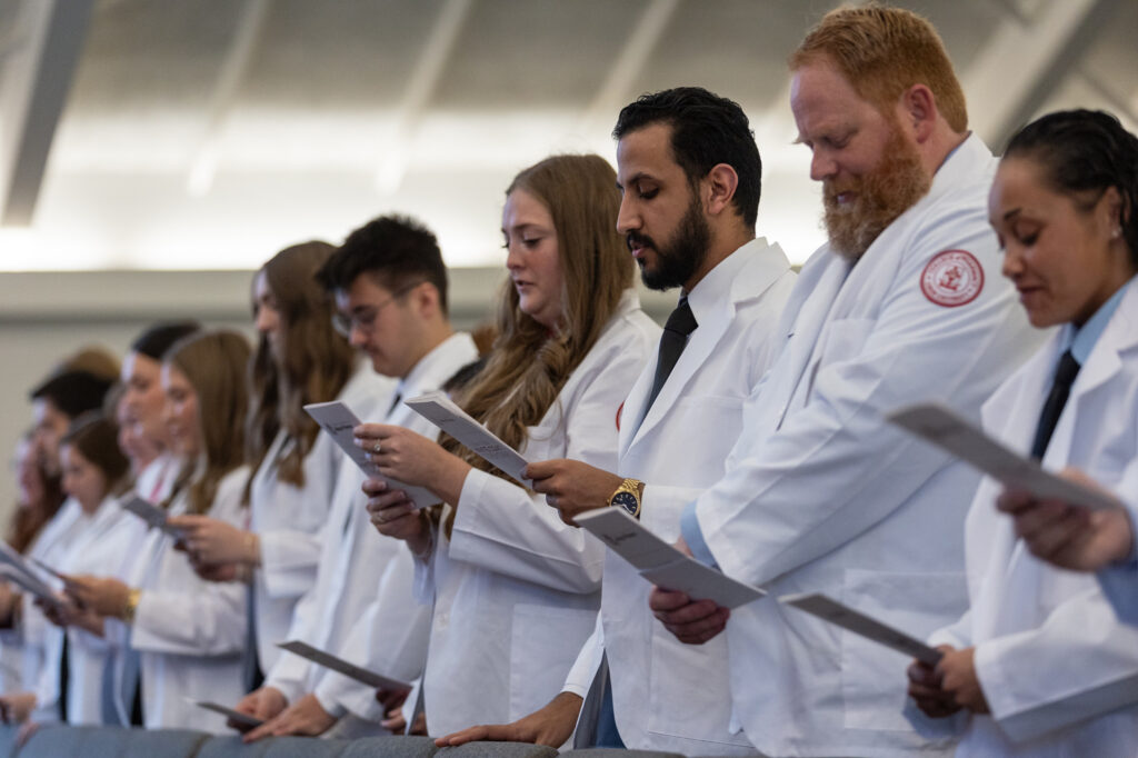 A group of people in white lab coats stand in a row indoors, holding papers and reading from them, likely participating in a PharmD ceremony or event.