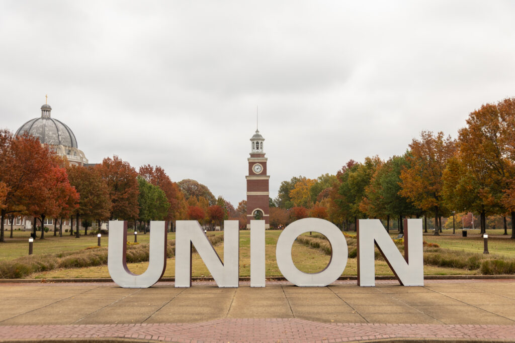 Large white letters spelling UNION stand on a sidewalk in front of autumn trees; Union's main campus is in Jackson, Tennessee. A domed building sits to the left, and a clock tower with a spire rises under the cloudy sky in the background.