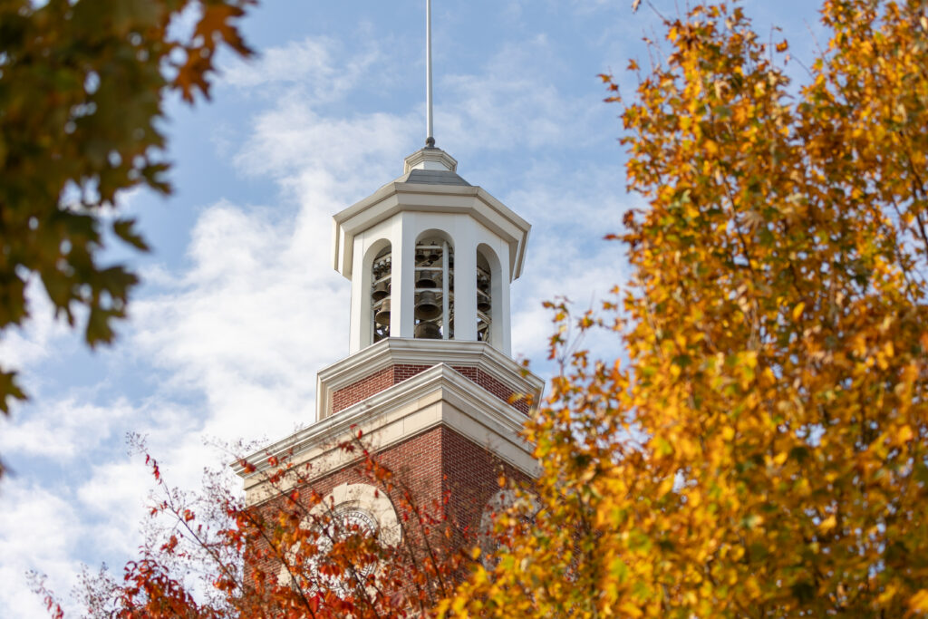 A brick clock tower with a white belfry rises above autumn trees with orange and yellow leaves on Union's main campus in Jackson, Tennessee, set against a blue sky with scattered clouds.