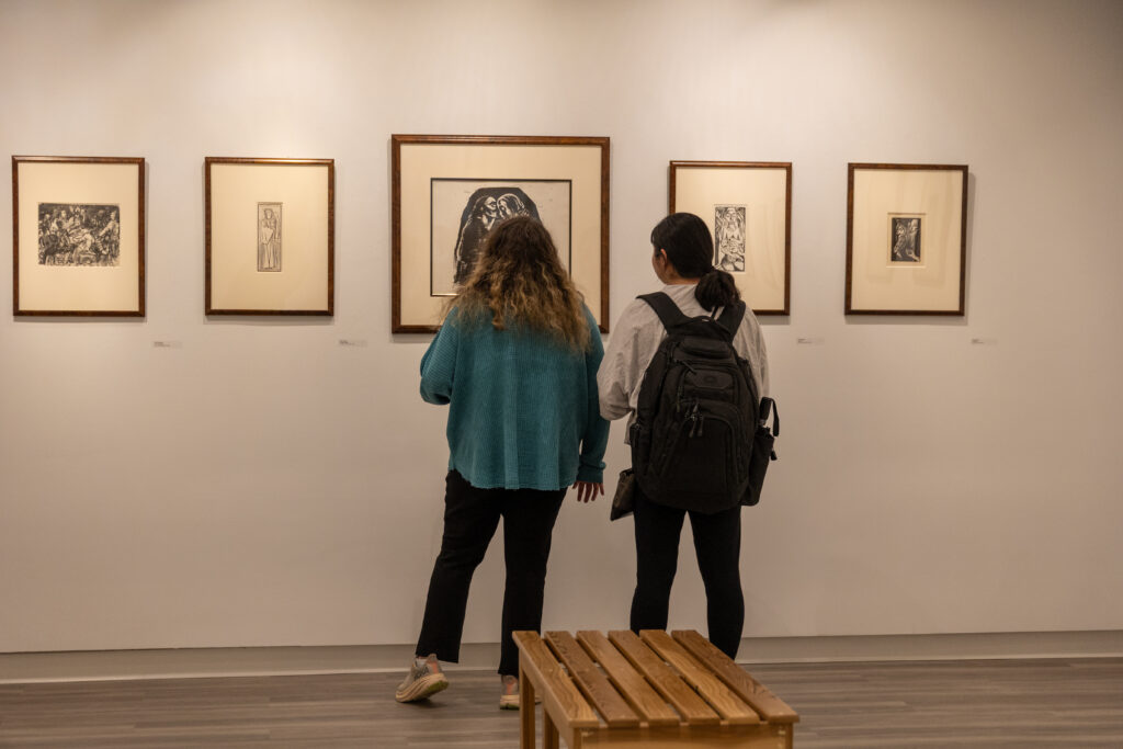 Two people stand in front of framed artwork on a gallery wall, observing the pieces. A wooden bench is in the foreground. The gallery has light-colored walls and hardwood floors.