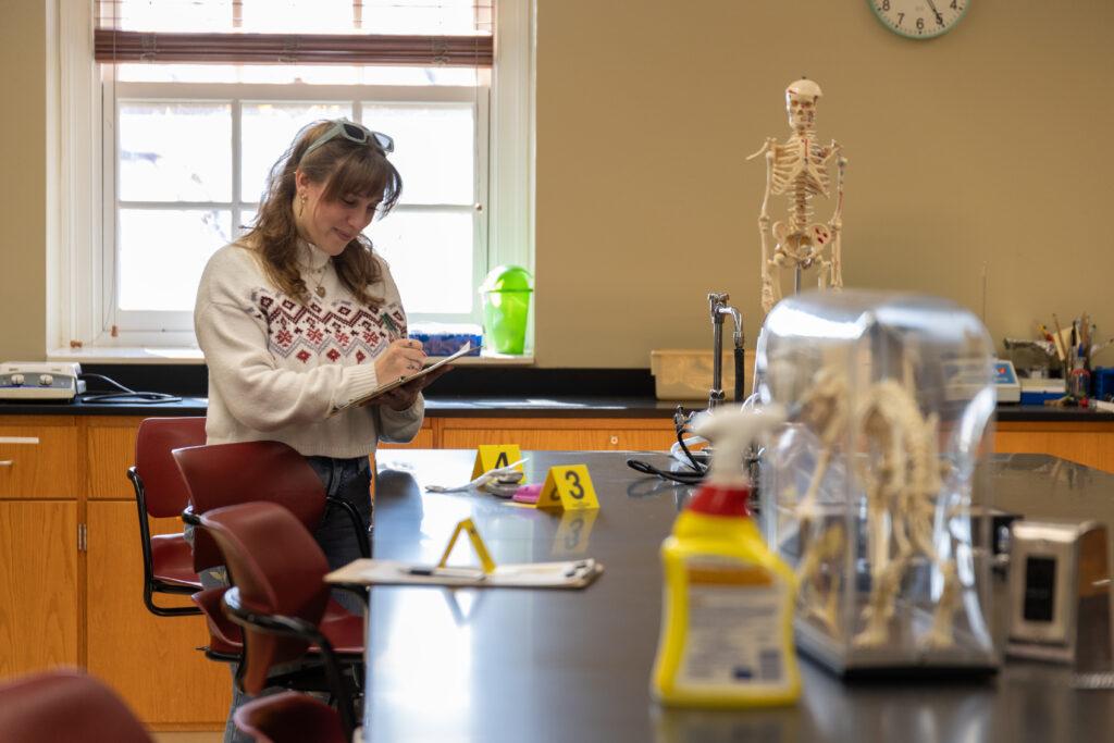 A person in a sweater writes on a clipboard beside a lab table marked with numbered evidence placards, a skeleton model, and cleaning supplies—capturing the investigative side of criminal justice—in a classroom with a window and clock.