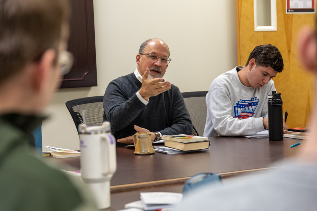 A man discusses classics and gestures while seated at a conference table with books and mugs in front of him, as another person beside him takes notes. Two more people are partially visible in the foreground.