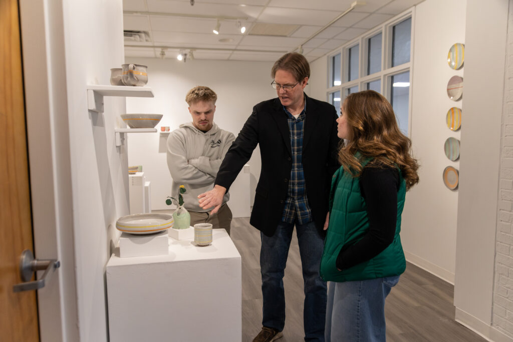 Three people stand in an art gallery, exploring the aesthetics of ceramic pieces displayed on white pedestals. One man gestures toward a vase, inviting discussion of visual theory as the others observe. Plates are mounted on the wall behind them.