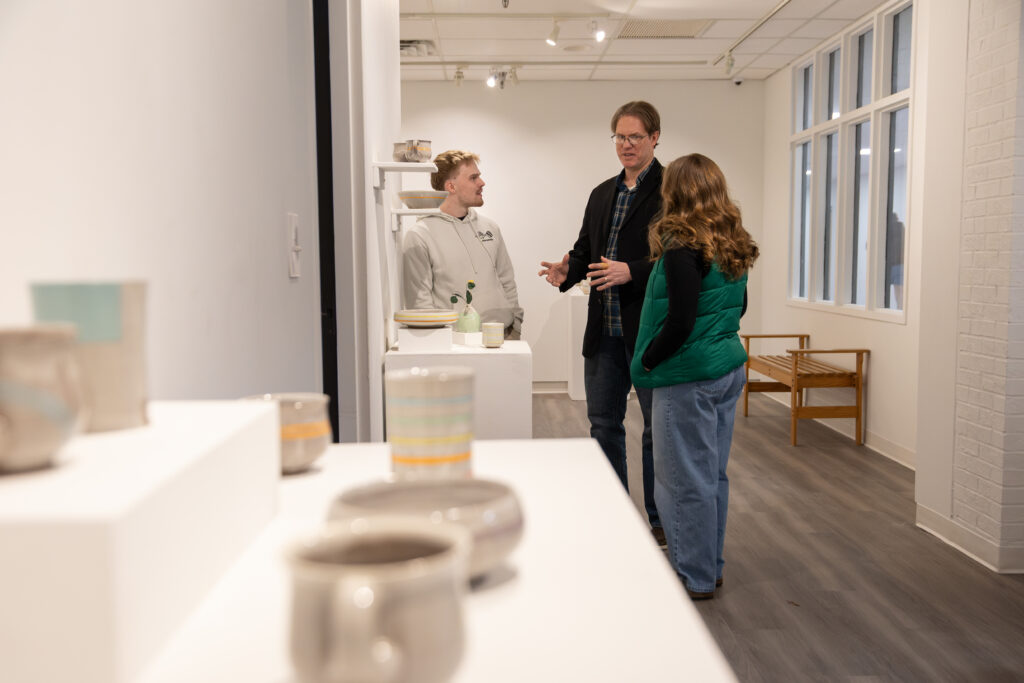 Three people stand and talk inside a modern art gallery with ceramic pieces displayed on white pedestals in the foreground. The gallery has white walls, large windows, and wood flooring.