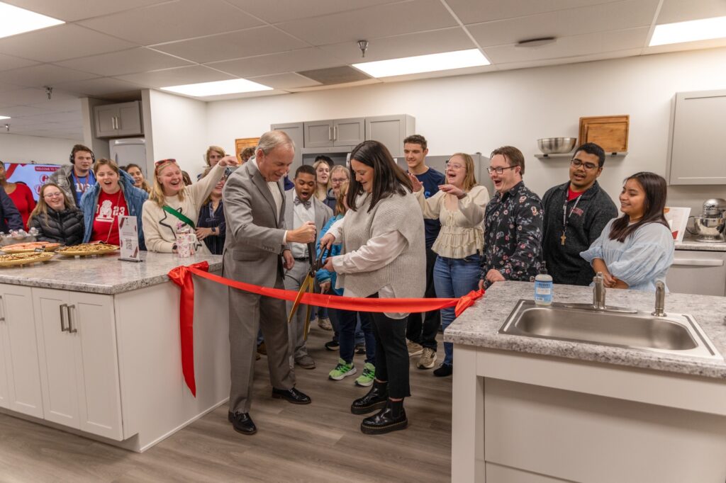 A man in a suit and a woman cut a red ribbon with large scissors in a kitchen, surrounded by smiling people. Countertops have food trays, and cabinets and appliances are visible in the background.