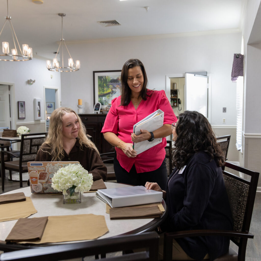 Three women hold a meeting at a round table in a brightly lit room. One woman stands holding paperwork and smiling, while the other two sit and listen. The table has folders, flowers, and a cardboard sign.