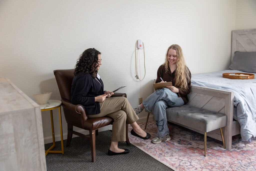 Two women sit across from each other in a bedroom. One, possibly a social work or human services professional, sits in an armchair with a clipboard, while the other writes on a notepad. Both appear engaged in conversation.