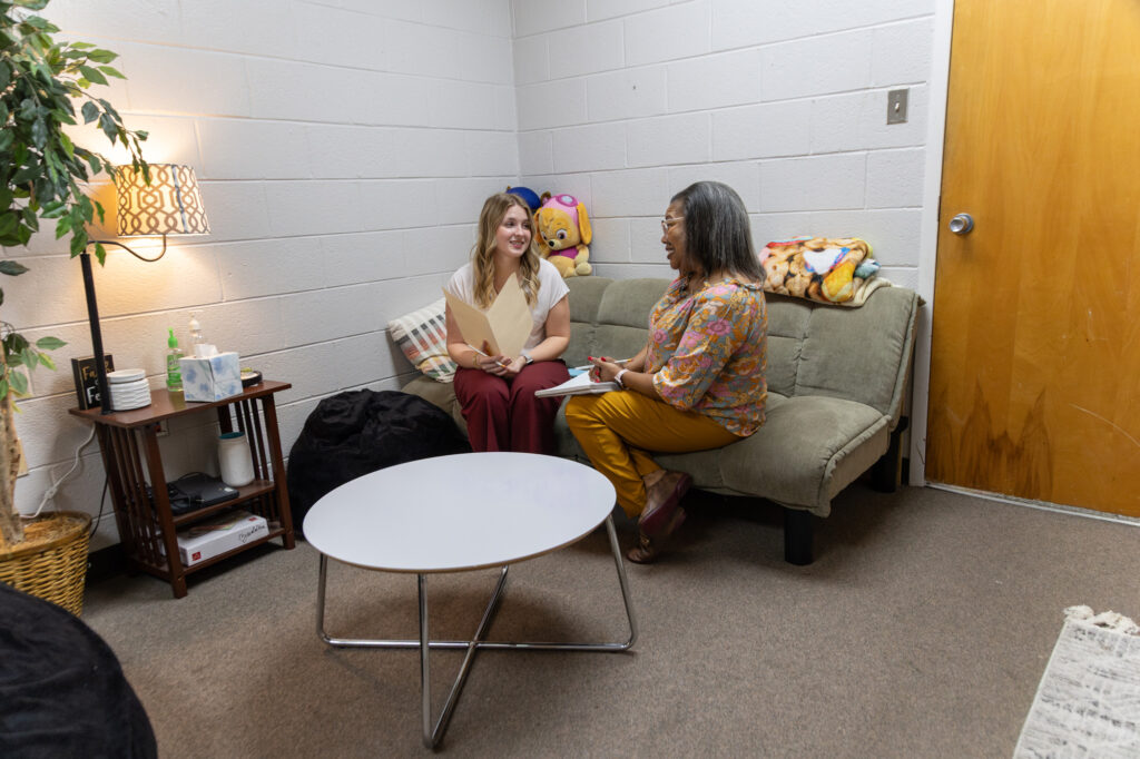 Two women sit on a small couch in an office, discussing social ministries. One holds a folder and smiles, while the other writes on a notepad. The room features a white brick wall, round table, lamp, plants, and a wooden door.