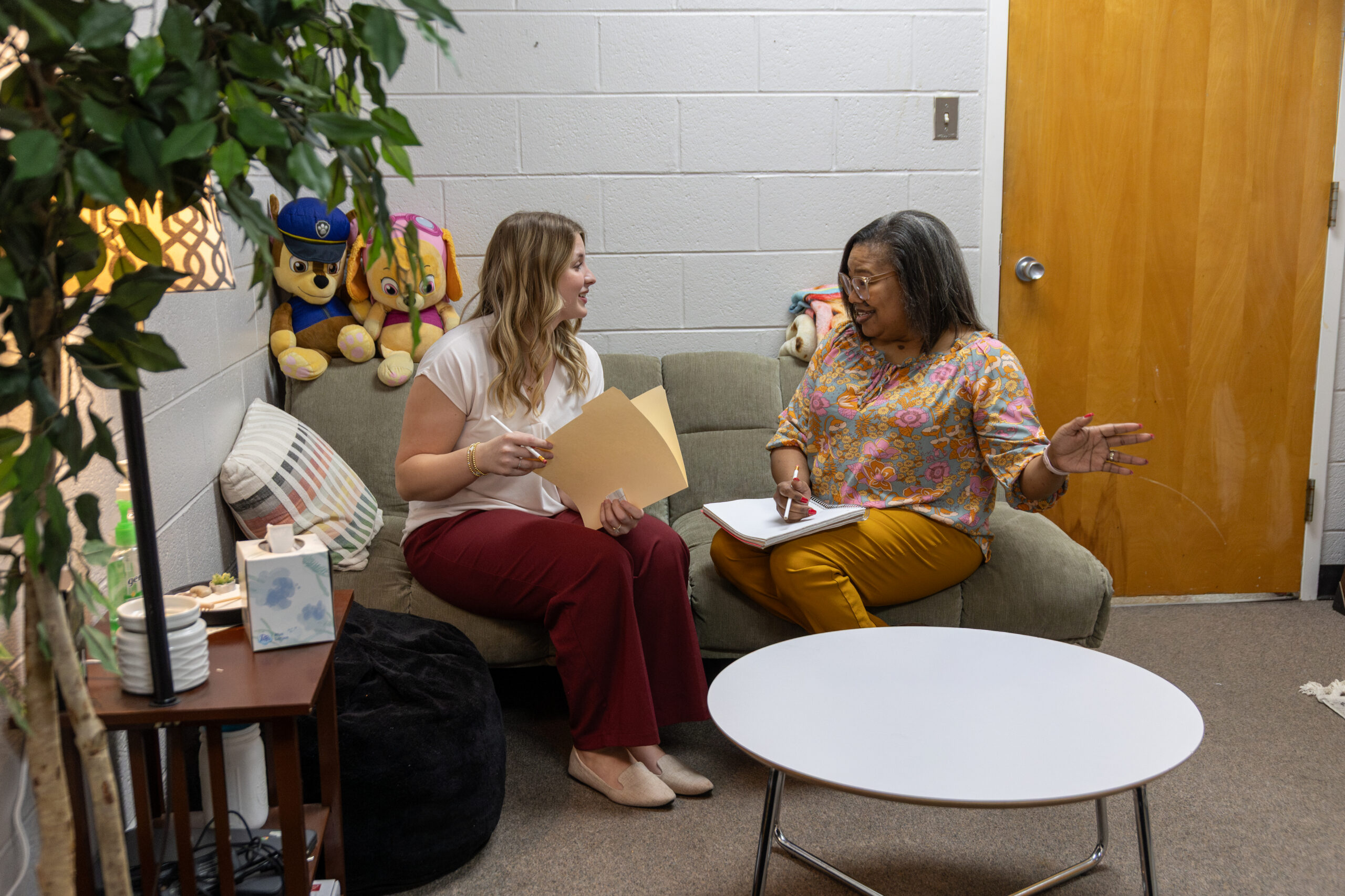 Two women, possibly discussing a Bachelor of Social Work program, sit on a couch in an office-like room with papers in hand. Stuffed toys rest behind them, with a small table in front and a wooden door to the side.