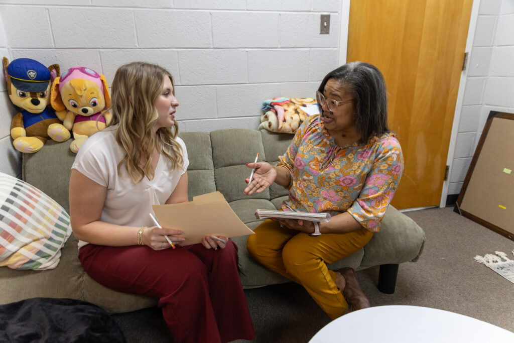 Two women sit on a couch in an office, talking and holding folders and notebooks. Stuffed animals are arranged on the back of the couch, adding comfort to this mental health support setting, while a blanket is draped nearby.