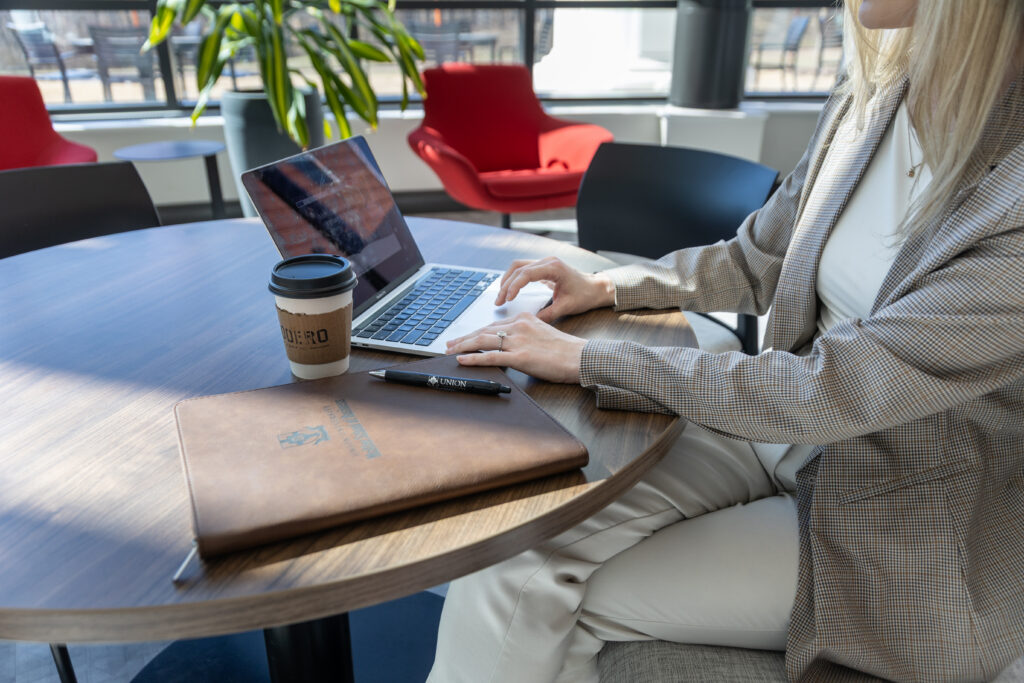A person in business attire works on a laptop at a round table, focusing on business administration tasks. A coffee cup, notebook, and pen are on the table. Bright chairs and windows are visible in the background.