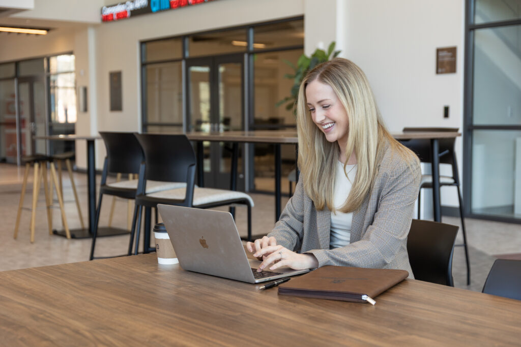 A woman with long blonde hair, wearing a blazer, sits at a table in a modern office space, typing on her laptop. Smiling at the screen, she appears to be working on business management tasks for her MBA studies.