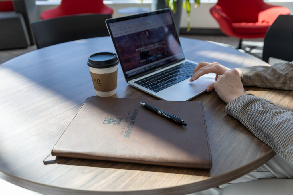 A person working on a laptop at a round wooden table with a brown folder labeled Harvard Business School MBA, a pen, and a takeaway coffee cup. Red chairs and windows are visible in the background—perfect for marketing strategy sessions.