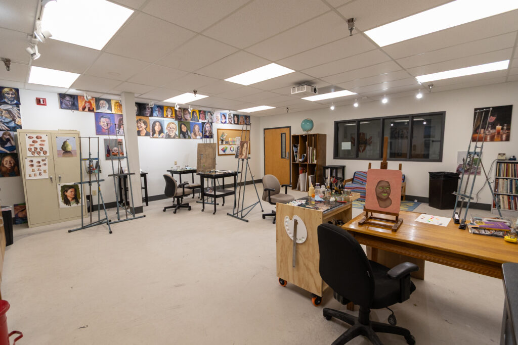 A spacious art classroom with easels, desks, stools, and art supplies. Various portraits and artwork are displayed on the walls. Large windows and overhead lights provide ample illumination.