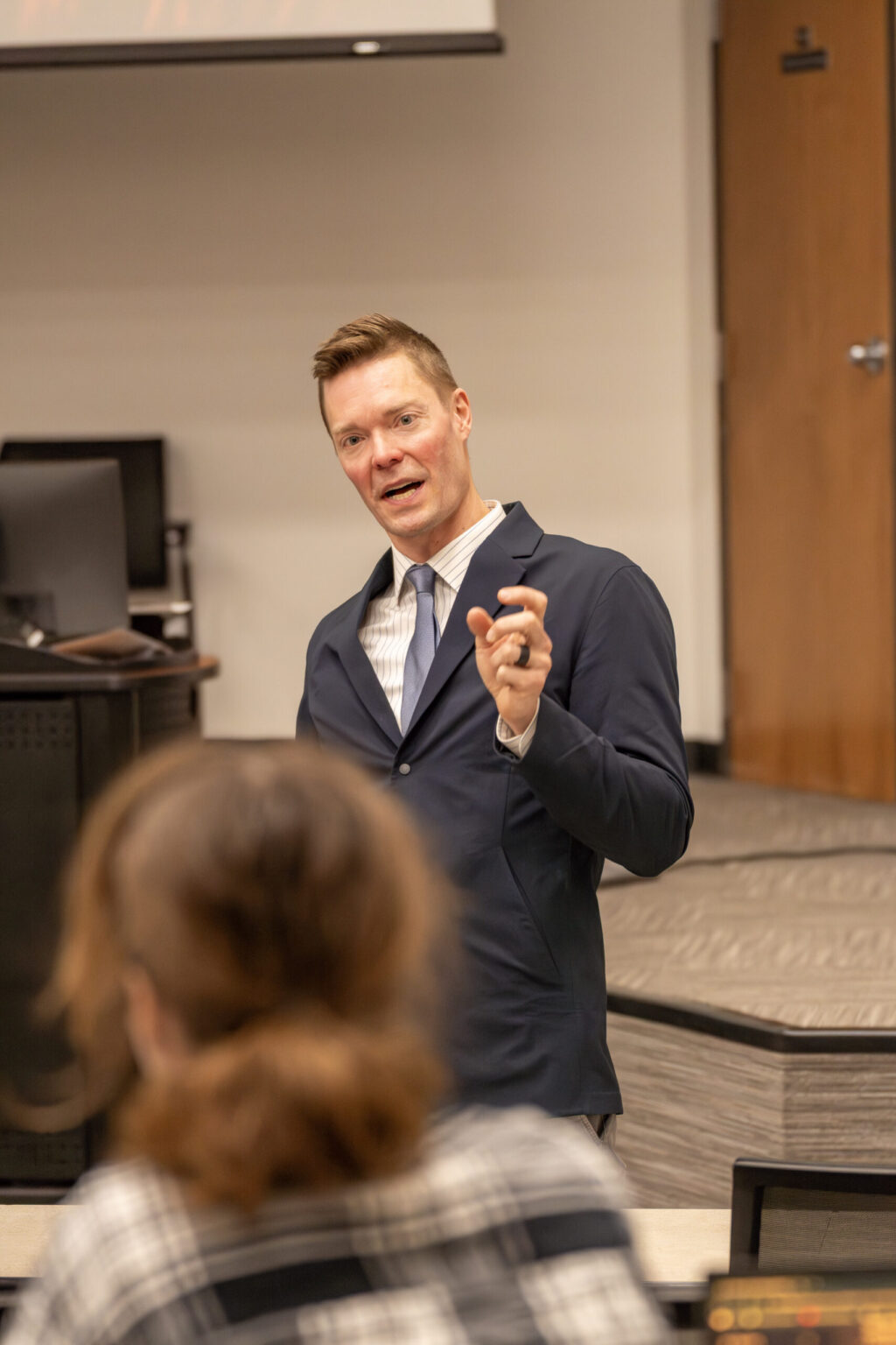 A man in a suit stands and gestures while speaking to a seated person in a classroom or lecture setting. There are computers and a wooden door in the background.
