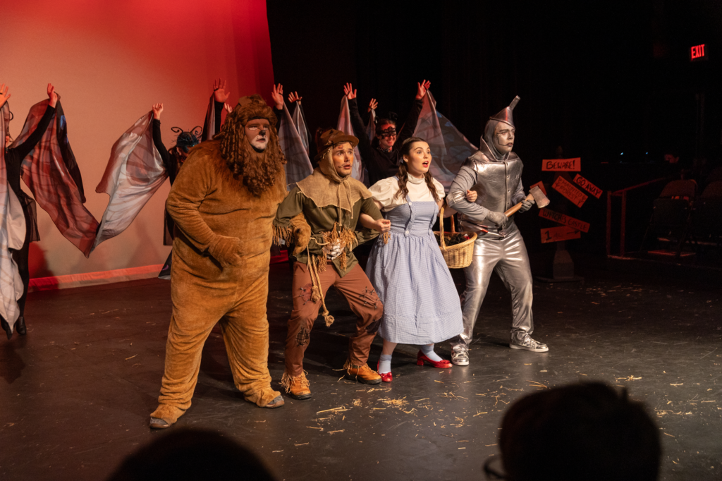 Four people dressed as the Cowardly Lion, Scarecrow, Dorothy, and Tin Man perform on stage in a lively music performance from The Wizard of Oz, with others in costume and colorful lighting in the background.