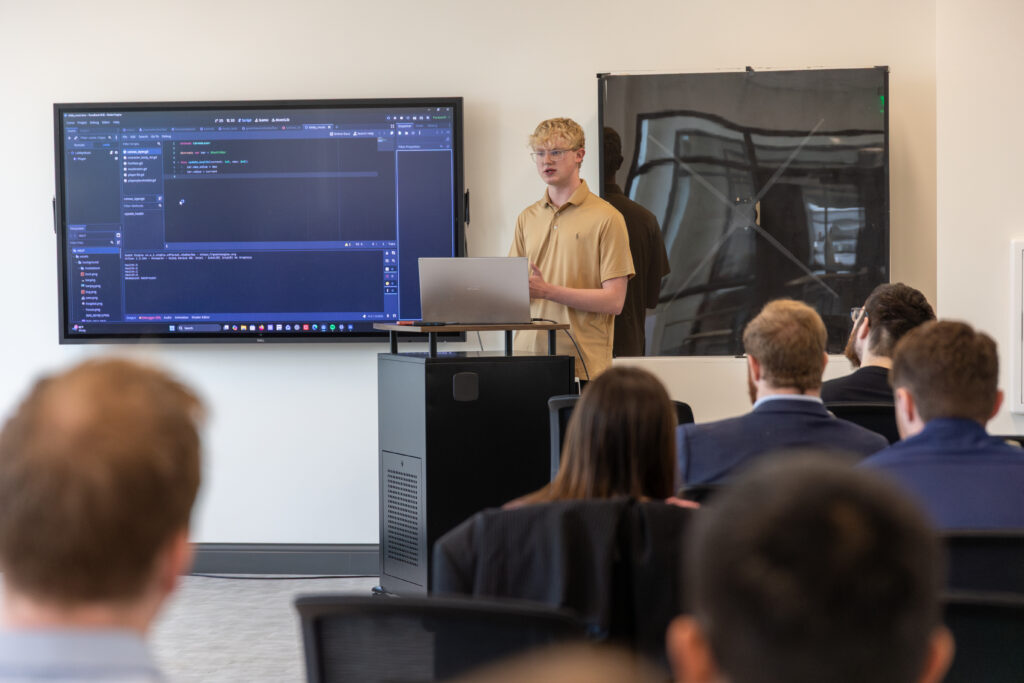 A person stands at a podium with a laptop, presenting game development coding information displayed on a large screen to an audience seated in a conference room.