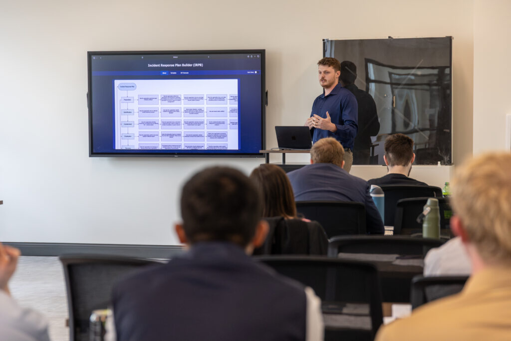 A man stands at the front of a classroom, presenting a slide titled Incident Response Plan Outline (IRPO) to an audience seated at desks. He uses his laptop to discuss key aspects of Information Systems in incident management.