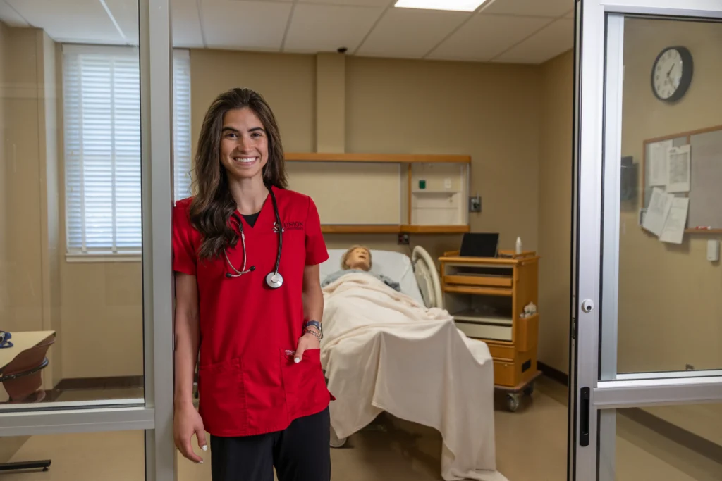 A person in red scrubs with a stethoscope smiles and stands near the entrance of a hospital room where a medical mannequin lies on a hospital bed, covered with a blanket.