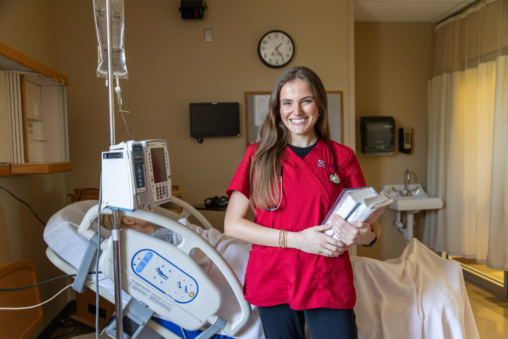 A woman in red medical scrubs stands in a hospital room, smiling and holding notebooks—ready for her Pre-Professional Health journey. Behind her is a hospital bed with medical equipment, a curtain, and a wall clock above the sink.
