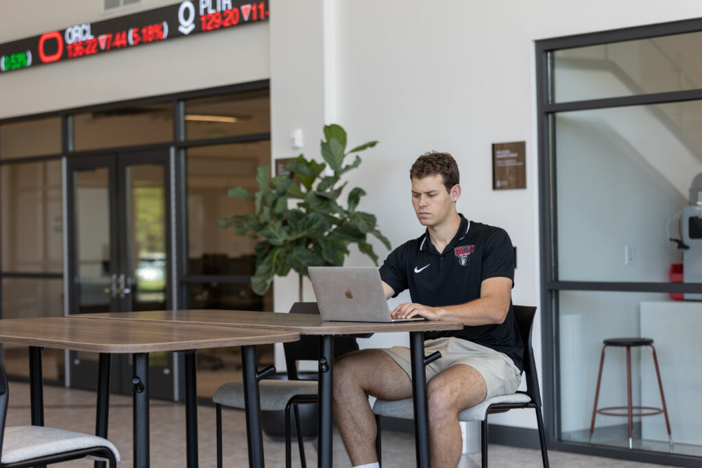 A young man wearing a black polo shirt and shorts sits at a table in a modern building, working on MBA business analytics assignments on his laptop. Digital stock tickers and glass doors are visible in the background.