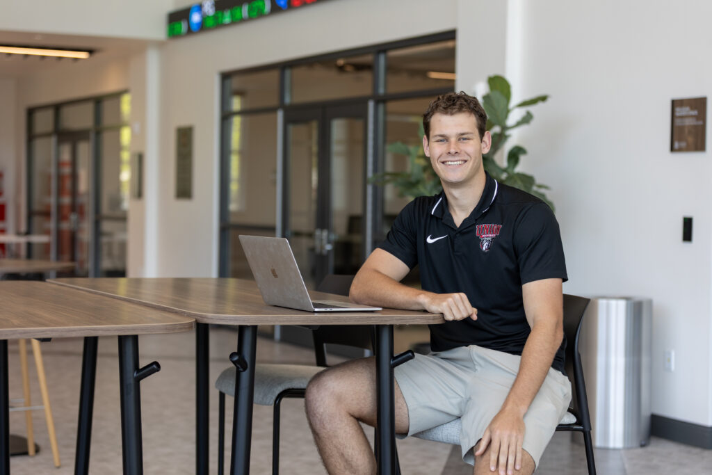 A young man in a black polo shirt and light shorts sits at a tall table in a modern indoor space, smiling at the camera with an open laptop in front of him.
